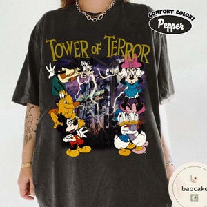 May include: Dark gray t-shirt featuring the text "TOWER OF TERROR" in yellow, with cartoon characters. The shirt has a distressed look and a "Comfort Colors Pepper" logo.