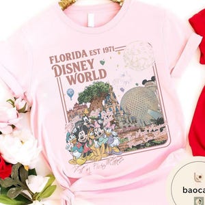 May include: A light pink short-sleeved t-shirt featuring a graphic print of Disney World landmarks and characters. The text reads Florida Est 1971 Disney World and The Most Magical Place On Earth, accompanied by illustrations of Mickey, Minnie, and friends.