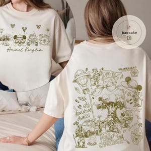May include: Cream-colored t-shirt with a green graphic design. The front features "Animal Kingdom" text and themed illustrations. The back displays "Adventure Awaits!" text, along with other illustrations and the text "Kilimanjaro".