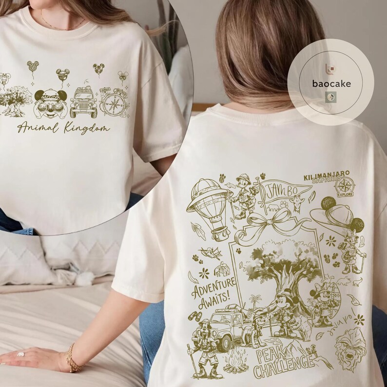 May include: Cream-colored t-shirt with a green graphic design. The front features "Animal Kingdom" text and themed illustrations. The back displays "Adventure Awaits!" text, along with other illustrations and the text "Kilimanjaro".