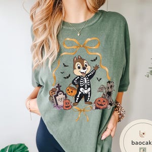 May include: Green t-shirt featuring a cartoon character in a skeleton costume, holding a pumpkin-shaped basket. The design includes Halloween elements like pumpkins, bats, and a tombstone. A gold ribbon frames the character.