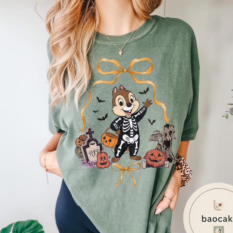 May include: Green t-shirt featuring a cartoon character in a skeleton costume, holding a pumpkin-shaped basket. The design includes Halloween elements like pumpkins, bats, and a tombstone. A gold ribbon frames the character.