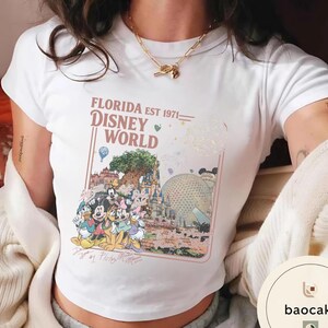 May include: A white short-sleeved t-shirt featuring a graphic print with the text FLORIDA EST 1971 DISNEY WORLD. The design includes illustrations of Disney characters, the Tree of Life, Cinderella Castle, and Spaceship Earth in a rectangular frame.