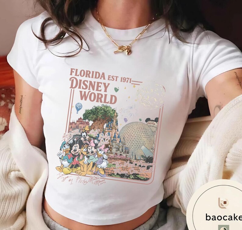 May include: A white short-sleeved t-shirt featuring a graphic print with the text FLORIDA EST 1971 DISNEY WORLD. The design includes illustrations of Disney characters, the Tree of Life, Cinderella Castle, and Spaceship Earth in a rectangular frame.