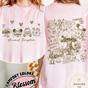 May include: A light pink t-shirt with a front graphic of Animal Kingdom icons and text. The back features a detailed illustration with the text "Adventure Awaits!" and "Peak Challenge". The shirt is labeled "Comfort Colors Blossom".
