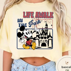 May include: A light yellow t-shirt with a graphic of Mickey Mouse and a castle. The text "LIFE A WALK IN THE Park" is printed above the graphic in red and blue.