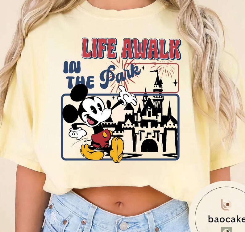 May include: A light yellow t-shirt with a graphic of Mickey Mouse and a castle. The text "LIFE A WALK IN THE Park" is printed above the graphic in red and blue.