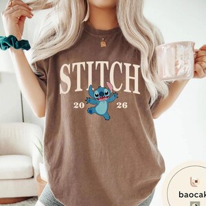 May include: Brown t-shirt featuring the word "STITCH" in large white letters, with a blue cartoon character and the numbers "20" and "26" below. The shirt is a neutral color.