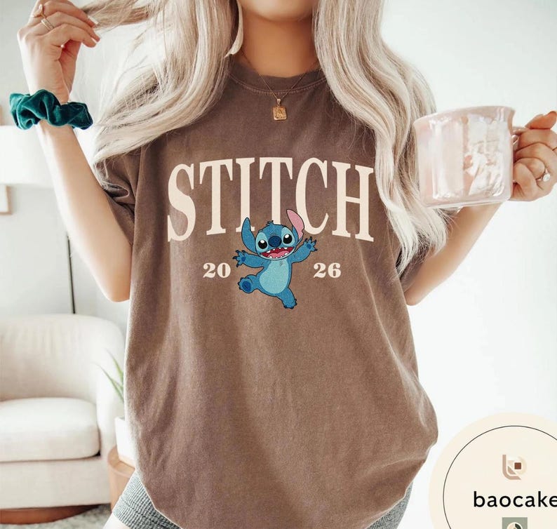 May include: Brown t-shirt featuring the word "STITCH" in large white letters, with a blue cartoon character and the numbers "20" and "26" below. The shirt is a neutral color.