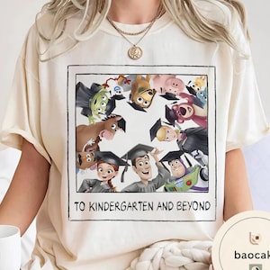 May include: A cream-coloured t-shirt with a framed graphic of Toy Story characters wearing graduation caps. The text "TO KINDERGARTEN AND BEYOND" is printed below the image. The shirt is a casual style.