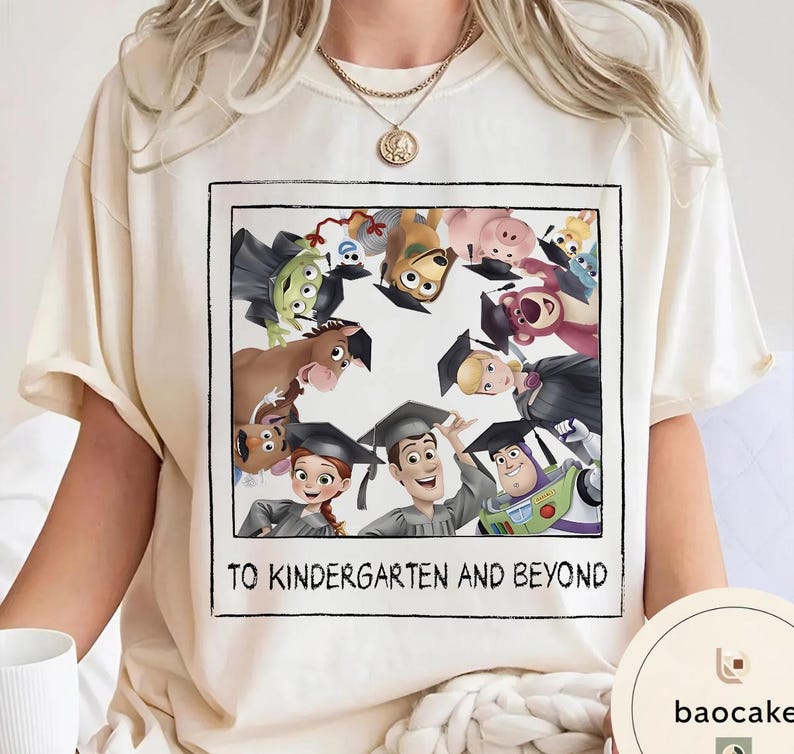 May include: A cream-coloured t-shirt with a framed graphic of Toy Story characters wearing graduation caps. The text "TO KINDERGARTEN AND BEYOND" is printed below the image. The shirt is a casual style.