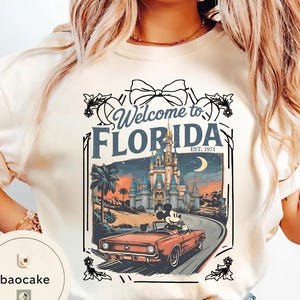 May include: Cream-colored t-shirt with a graphic that says "Welcome to Florida" and features a vintage illustration of Mickey Mouse driving a red convertible towards a castle. The design includes palm trees, a crescent moon, and the text "EST. 1971".