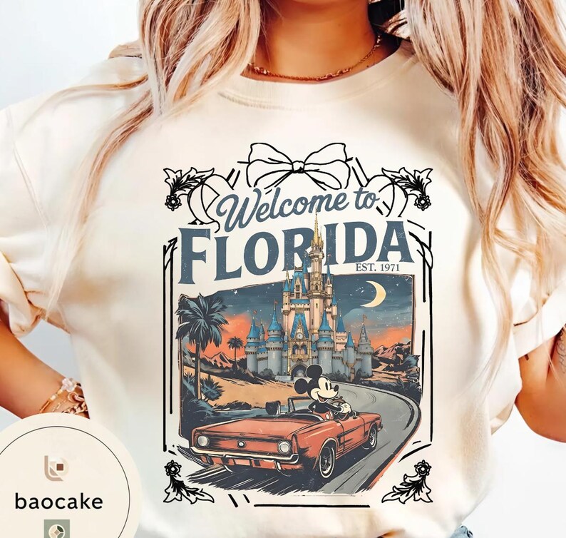 May include: Cream-colored t-shirt with a graphic that says "Welcome to Florida" and features a vintage illustration of Mickey Mouse driving a red convertible towards a castle. The design includes palm trees, a crescent moon, and the text "EST. 1971".