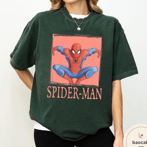 May include: Dark green t-shirt featuring a graphic of Spider-Man in a crouched pose within a coral-colored rectangle. The words "SPIDER-MAN" are printed below the image in a matching coral color. The shirt has short sleeves and a crew neck.