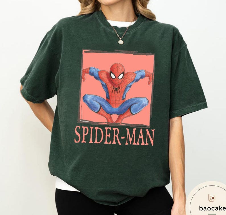 May include: Dark green t-shirt featuring a graphic of Spider-Man in a crouched pose within a coral-colored rectangle. The words "SPIDER-MAN" are printed below the image in a matching coral color. The shirt has short sleeves and a crew neck.