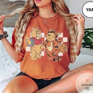 May include: Burnt orange t-shirt featuring a cartoon illustration of Winnie the Pooh and Tigger. Winnie the Pooh is holding a blue bag and blowing a bubble. Tigger is holding a pumpkin. The background is a white and red checkered pattern.