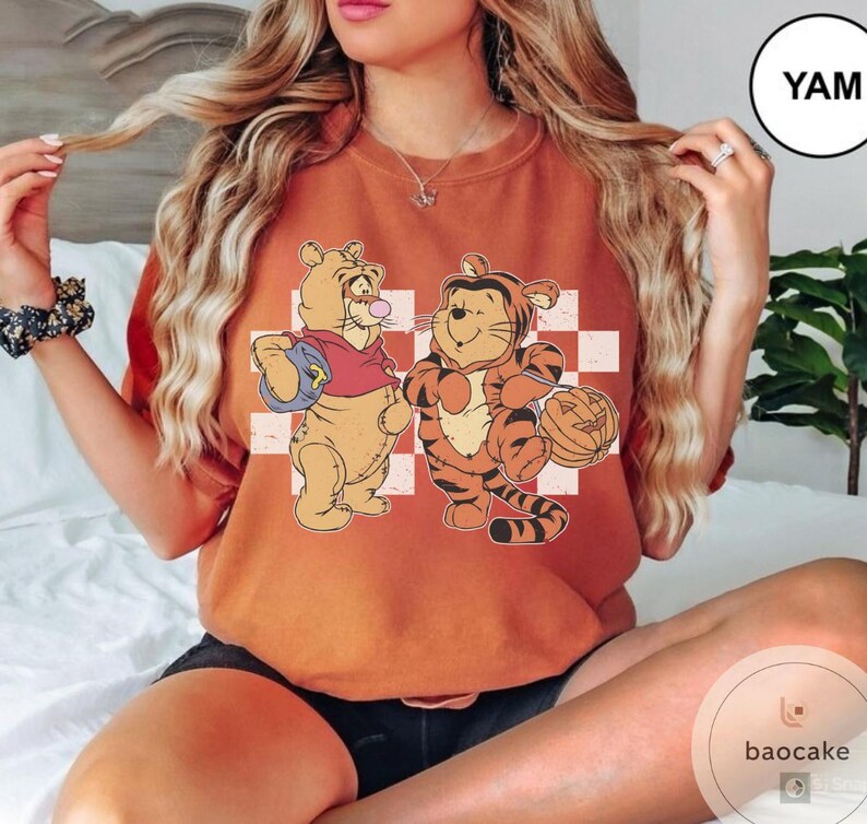 May include: Burnt orange t-shirt featuring a cartoon illustration of Winnie the Pooh and Tigger. Winnie the Pooh is holding a blue bag and blowing a bubble. Tigger is holding a pumpkin. The background is a white and red checkered pattern.