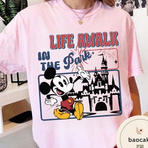 May include: Pale pink t-shirt with a graphic of Mickey Mouse pointing towards a castle, with the text "LIFE AWALK IN THE Park". The design includes fireworks and a blue border. The shirt is a light pink colour.