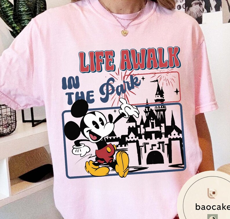 May include: Pale pink t-shirt with a graphic of Mickey Mouse pointing towards a castle, with the text "LIFE AWALK IN THE Park". The design includes fireworks and a blue border. The shirt is a light pink colour.