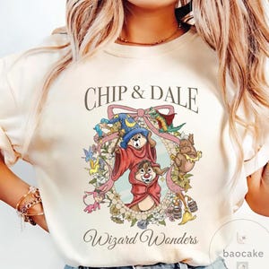 May include: Cream-colored sweatshirt featuring Chip and Dale in wizard attire, surrounded by a floral wreath and a pink ribbon. The text "Chip & Dale" and "Wizard Wonders" are displayed above and below the graphic.
