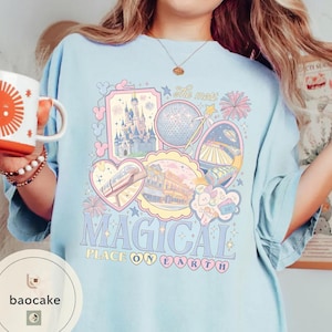 May include: Pale blue t-shirt with a colourful graphic design featuring a castle, a globe, and the text "MAGICAL PLACE ON EARTH". The design includes pastel pink, blue, and yellow elements. The shirt has short sleeves and a crew neck.
