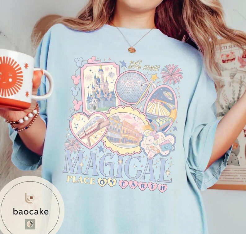 May include: Pale blue t-shirt with a colourful graphic design featuring a castle, a globe, and the text "MAGICAL PLACE ON EARTH". The design includes pastel pink, blue, and yellow elements. The shirt has short sleeves and a crew neck.