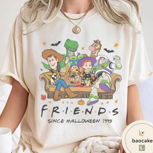 May include: A cream-coloured t-shirt featuring a graphic of Toy Story characters sitting on a couch. The text reads "F R I E N D S" and "SINCE HALLOWEEN 1995". The design includes autumn leaves, bats, and pumpkins, with characters like Woody, Buzz Lightyear, and Jessie.
