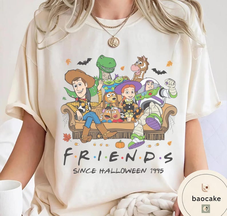 May include: A cream-coloured t-shirt featuring a graphic of Toy Story characters sitting on a couch. The text reads "F R I E N D S" and "SINCE HALLOWEEN 1995". The design includes autumn leaves, bats, and pumpkins, with characters like Woody, Buzz Lightyear, and Jessie.