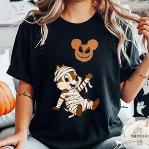 May include: Black t-shirt featuring a cartoon chipmunk wrapped in mummy bandages, with a jack-o'-lantern balloon. The chipmunk is brown and white, with a smiling expression. The balloon is a brown Mickey Mouse head with a carved face.