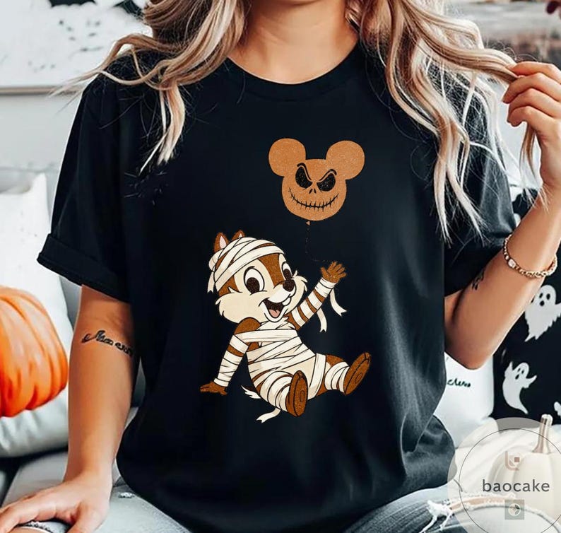 May include: Black t-shirt featuring a cartoon chipmunk wrapped in mummy bandages, with a jack-o'-lantern balloon. The chipmunk is brown and white, with a smiling expression. The balloon is a brown Mickey Mouse head with a carved face.