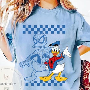 May include: Light blue t-shirt featuring a cartoon illustration of Donald Duck dressed as Spider-Man. The design includes a blue and white checkered pattern and a Spider-Man outline. The shirt has a crew neck.