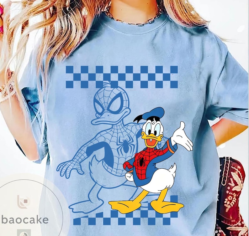 May include: Light blue t-shirt featuring a cartoon illustration of Donald Duck dressed as Spider-Man. The design includes a blue and white checkered pattern and a Spider-Man outline. The shirt has a crew neck.