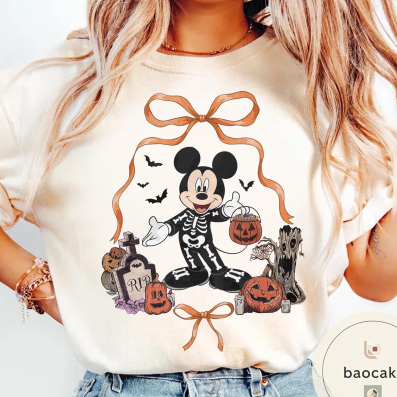 May include: Cream-colored t-shirt featuring a Halloween-themed Mickey Mouse design. Mickey is dressed as a skeleton, holding a jack-o'-lantern. The design includes pumpkins, bats, a tombstone, and decorative bows, creating a festive and spooky aesthetic.