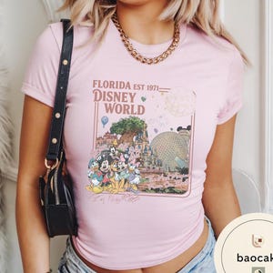 May include: A light pink short-sleeve t-shirt featuring a graphic print of Disney World landmarks and characters. The text reads FLORIDA EST 1971 DISNEY WORLD and The Most Magical Place On Earth, with illustrations of Mickey, Minnie, Donald, and Pluto.