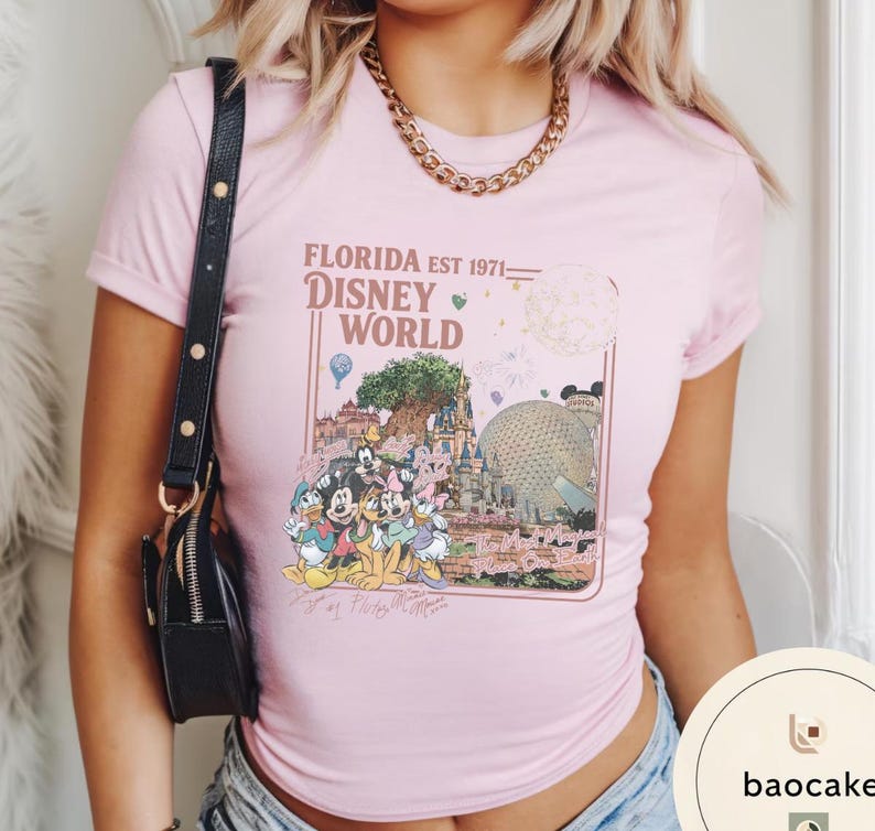 May include: A light pink short-sleeve t-shirt featuring a graphic print of Disney World landmarks and characters. The text reads FLORIDA EST 1971 DISNEY WORLD and The Most Magical Place On Earth, with illustrations of Mickey, Minnie, Donald, and Pluto.