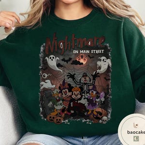 May include: Dark green sweatshirt with a Halloween-themed graphic. The graphic features the text "Nightmare on Main Street" with cartoon characters dressed in Halloween costumes, including ghosts, bats, and pumpkins. The design has a spooky, festive vibe.