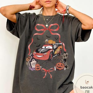 May include: Dark gray t-shirt featuring a Halloween-themed graphic. The design includes a red race car with a skeleton design, pumpkins, bats, and a spider web. The number 95 is visible on the car.