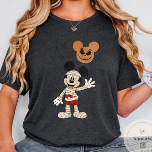 May include: Dark gray t-shirt featuring a Halloween-themed graphic. The design includes a mummy-style Mickey Mouse character and a Jack Skellington-inspired Mickey head. The shirt is a short-sleeved crew neck.