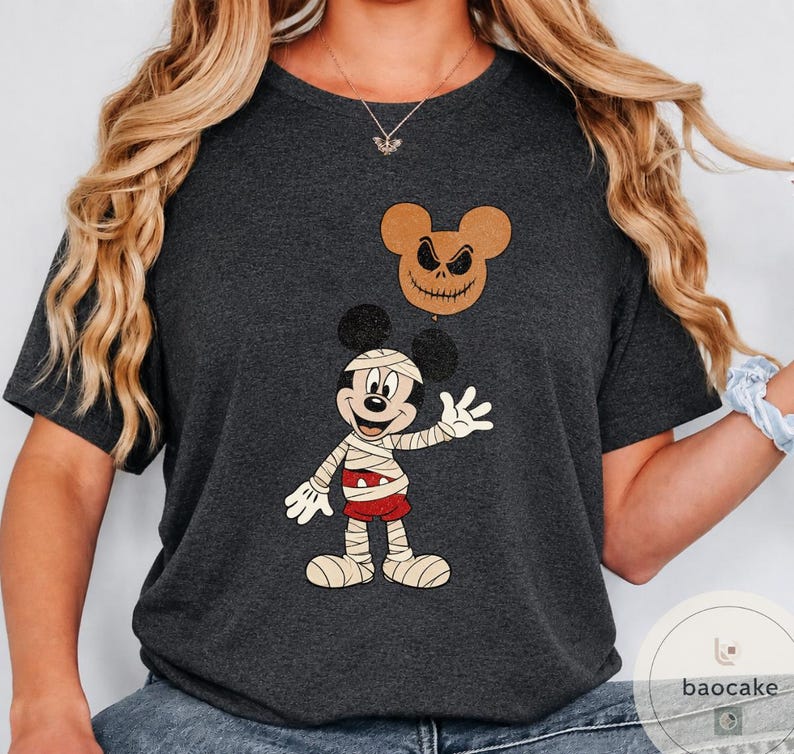 May include: Dark gray t-shirt featuring a Halloween-themed graphic. The design includes a mummy-style Mickey Mouse character and a Jack Skellington-inspired Mickey head. The shirt is a short-sleeved crew neck.