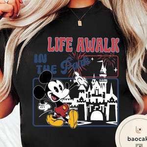 May include: Black t-shirt with a graphic of Mickey Mouse and the text "LIFE AWALK IN THE Park". The design includes a fireworks display and a castle silhouette. Mickey is wearing red shorts and yellow shoes, all within a rectangular frame.