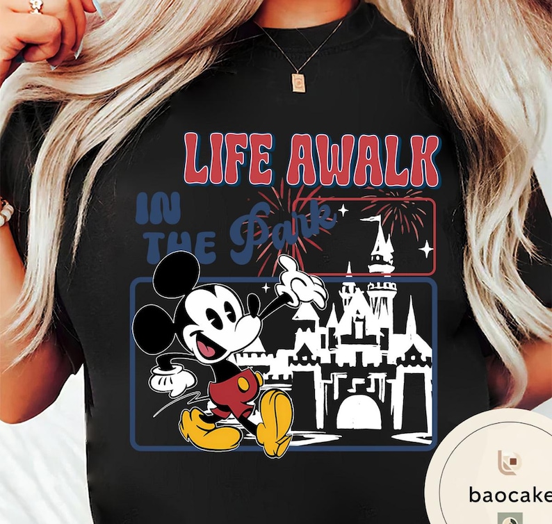 May include: Black t-shirt with a graphic of Mickey Mouse and the text "LIFE AWALK IN THE Park". The design includes a fireworks display and a castle silhouette. Mickey is wearing red shorts and yellow shoes, all within a rectangular frame.