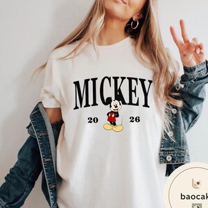 May include: Cream-colored t-shirt featuring the word "MICKEY" in bold black letters above a cartoon image of Mickey Mouse. The numbers "20" and "26" flank the character. The shirt is part of a clothing collection.