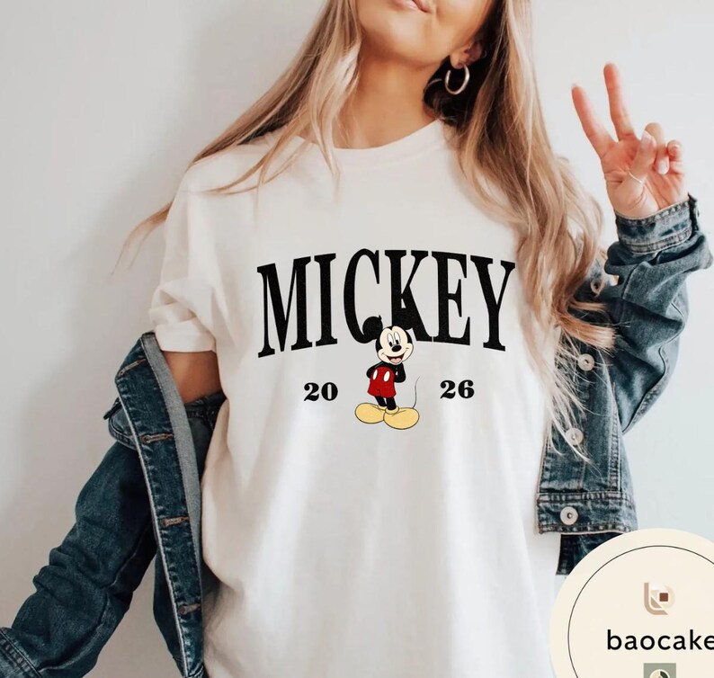 May include: Cream-colored t-shirt featuring the word "MICKEY" in bold black letters above a cartoon image of Mickey Mouse. The numbers "20" and "26" flank the character. The shirt is part of a clothing collection.