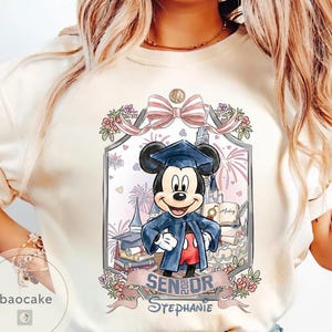 May include: Cream-colored t-shirt featuring a graduation-themed Mickey Mouse design. Mickey is wearing a graduation cap and gown, with a banner that reads "Senior Stephanie". The design includes a castle, fireworks, and floral accents.