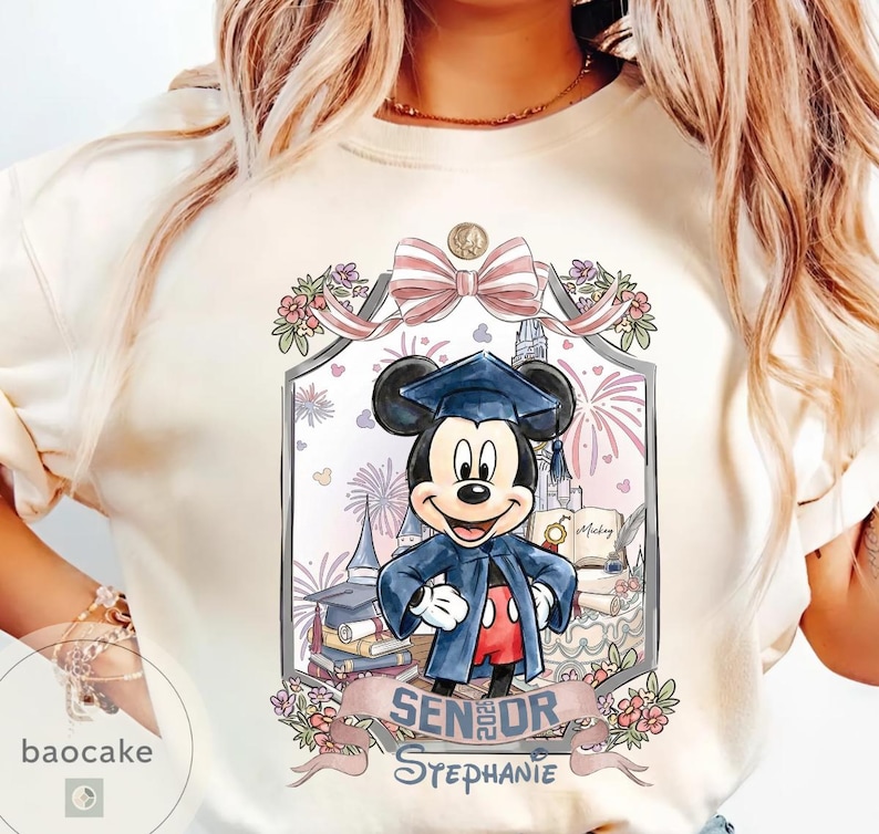 May include: Cream-colored t-shirt featuring a graduation-themed Mickey Mouse design. Mickey is wearing a graduation cap and gown, with a banner that reads "Senior Stephanie". The design includes a castle, fireworks, and floral accents.