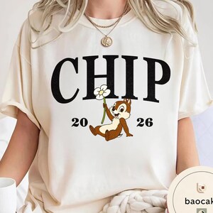 May include: Cream-colored t-shirt featuring the word "CHIP" in large black letters above a cartoon chipmunk holding a flower. The numbers "20" and "26" flank the chipmunk. The shirt has short sleeves.