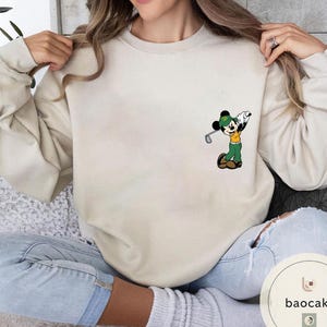 May include: Cream-colored sweatshirt featuring a cartoon Mickey Mouse playing golf. Mickey is wearing a green hat, yellow shirt, and green pants. The sweatshirt is a neutral color, suitable for casual wear. The design is on the left chest area.