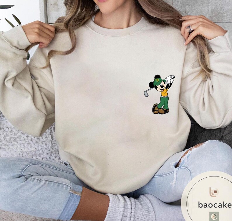 May include: Cream-colored sweatshirt featuring a cartoon Mickey Mouse playing golf. Mickey is wearing a green hat, yellow shirt, and green pants. The sweatshirt is a neutral color, suitable for casual wear. The design is on the left chest area.