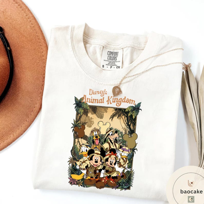 May include: Cream-colored t-shirt featuring a graphic of Disney's Animal Kingdom characters, including Mickey and Minnie Mouse, Donald Duck, and Goofy. The design is framed by jungle foliage. A gold necklace and a brown hat are also visible.