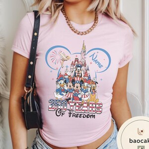 Peut inclure: T-shirt rose clair avec un motif de personnages Disney en tenues patriotiques devant un ch&acirc;teau. Le t-shirt porte l'inscription "250 Years of Freedom" et un espace pour un nom dans un contour d'oreille de Mickey.